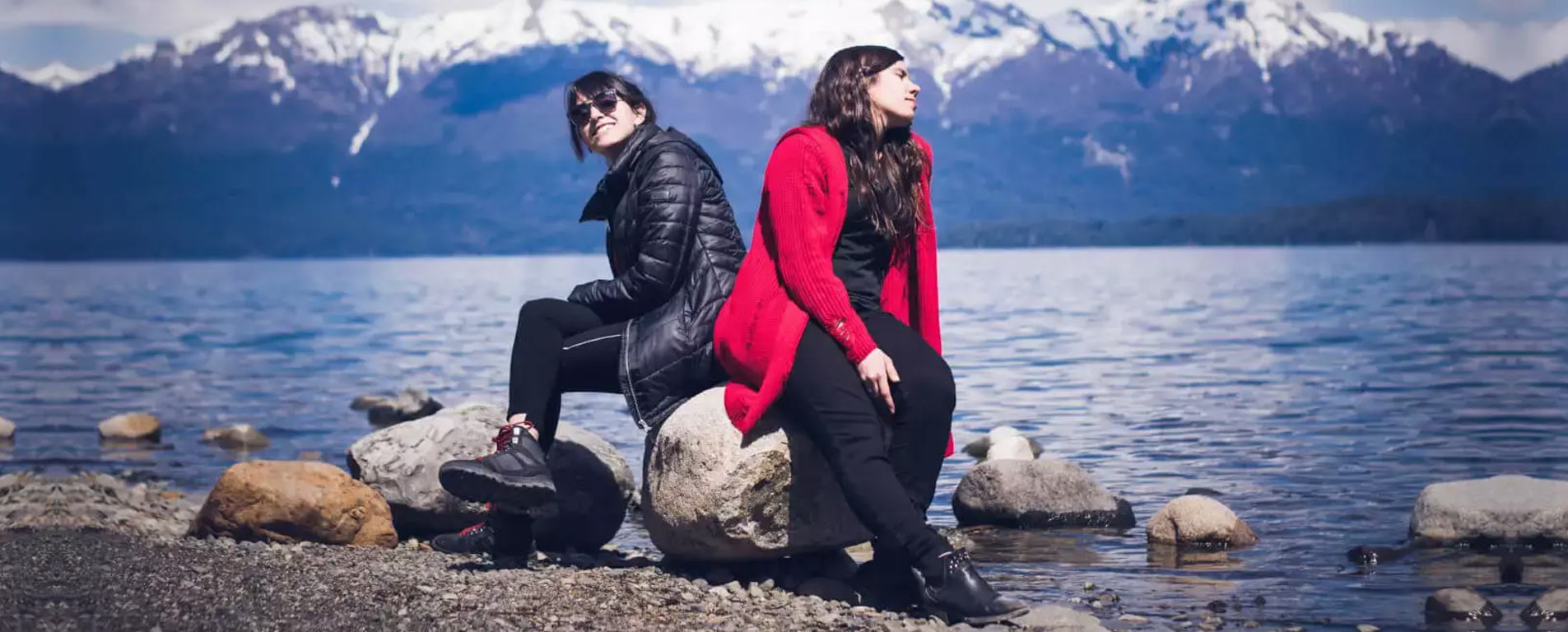 two women sitting on a rock in front of a lake